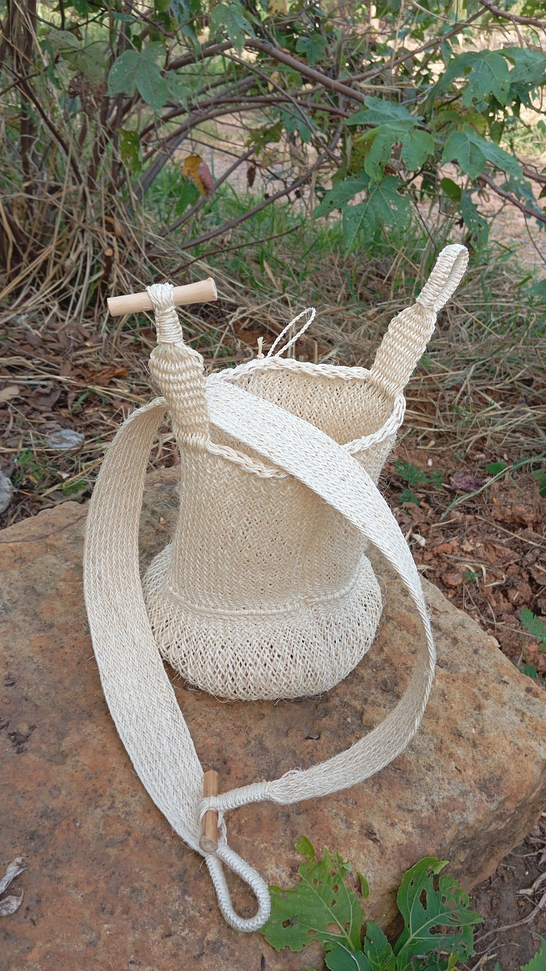 Mochila de fique tejida a mano con asa de madera, sobre una piedra en el campo